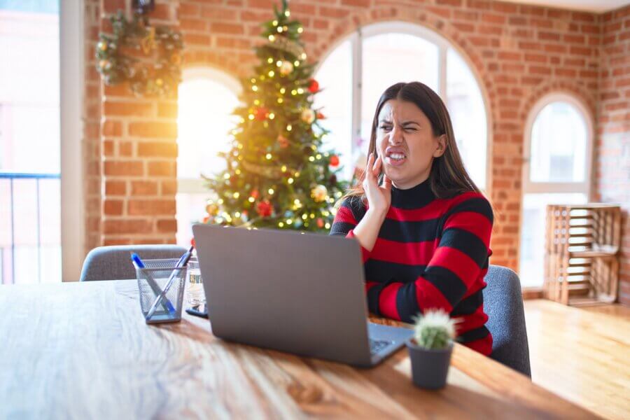 Beautiful woman sitting at the table working with laptop at home around christmas tree touching mouth with hand with painful expression because of toothache or dental illness on teeth. Dentist concept.